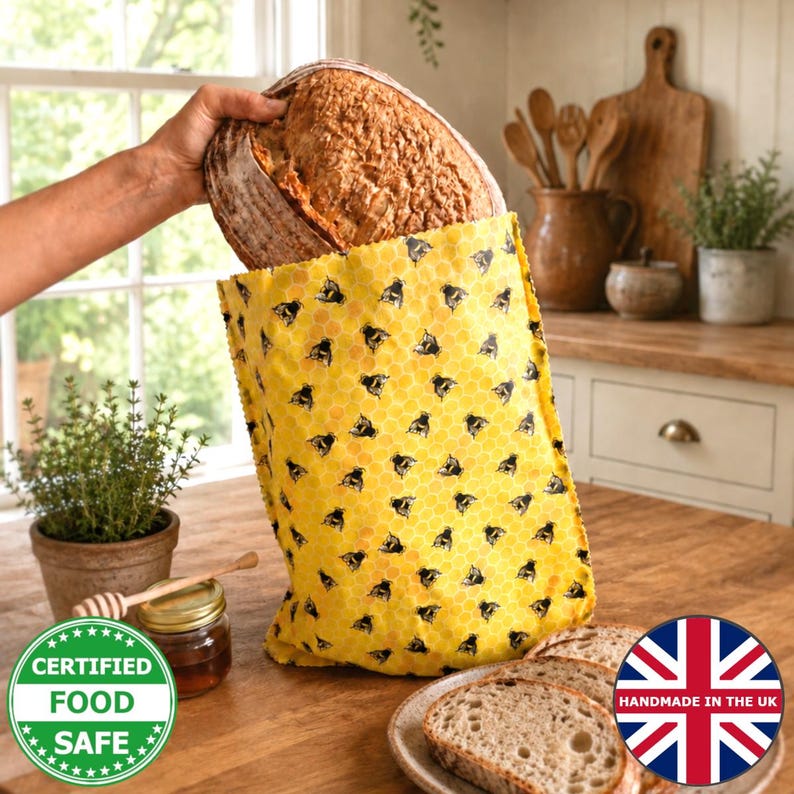 May include: A yellow beeswax food wrap bag featuring a repeating pattern of black bees and a honeycomb design. The bag is filled with a round loaf of crusty sourdough bread. The fabric wrap is flexible and designed for food storage.