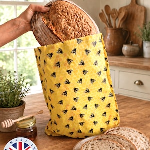 May include: A loaf of bread is held inside a yellow bread bag with a bee pattern. The bag is on a wooden surface with a jar of honey, a honey dipper, and a plate with sliced bread. The bag has a "100% Handmade" sticker.