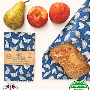 May include: A still-life image featuring a pear, two red apples, and blue beeswax food wraps. The wraps, patterned with white chickens, are shown folded and wrapped around a loaf of bread. The text on the wrap reads "Honey Bee Good Beeswax Food Wraps".