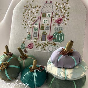 Può includere: Una collezione di zucche di stoffa fatte a mano in tonalità di verde e viola, con steli marroni e dettagli in filo di rame. Sullo sfondo un disegno a punto croce di una casa e uccelli. Il testo "mylanney" e "xxx" è visibile.