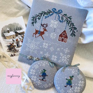 Può includere: Una scena natalizia a punto croce blu chiaro con una renna, una casa di pan di zenzero e fiocchi di neve. Sono presenti anche due ornamenti rotondi con sciatori e fiocchi di neve. Una cartolina vintage invernale e un nastro completano il tema.