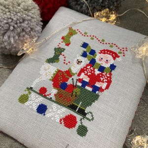 Può includere: Un ricamo a punto croce con una figura di Babbo Natale che tiene un sacco di regali, con un cane bianco seduto accanto a lui. Il disegno è su un tessuto grigio con uno sfondo bianco e include il testo "myfanny".