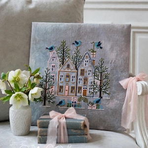May include: A square gray linen fabric panel featuring a cross-stitch embroidery of three houses, evergreen trees, and blue birds. The design includes small gift boxes and is accented with beads. The embroidery is displayed on a gray surface.