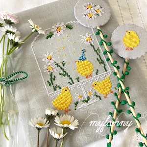 Op de afbeelding: Een kruissteekontwerp met drie gele kuikens en madeliefjes. De kuikens staan in een vierkant kader, één met een blauwe feesthoed. Twee ronde kruissteekontwerpen op stokjes tonen madeliefjes en een kuiken.