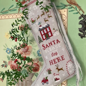 Può includere: Una calza di Natale grigia con disegni a punto croce. La calza presenta una casa rossa, Babbo Natale su una slitta, renne e il testo "SANTA PLEASE stop HERE". Ulteriori disegni includono un pupazzo di neve, regali e un albero di Natale.