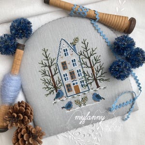 Può includere: Un ricamo a punto croce con una scena invernale con una casa, alberi e uccelli. Il disegno è su un tessuto grigio chiaro, con accenti blu e marroni. Pompon blu decorativi, rocchetti e pigne sono disposti attorno al ricamo.