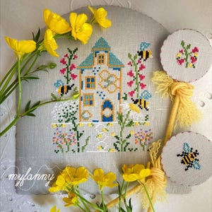 Può includere: Un disegno a punto croce con una casa dal tetto blu, circondata da fiori e api. Sono presenti anche due piccoli pezzi rotondi a punto croce con disegni floreali e di api. Fiori gialli e il testo "myfanny" sono visibili.