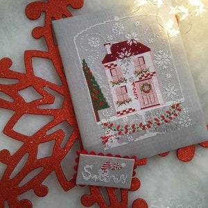 Può includere: Un ricamo a punto croce di una casa innevata con un tetto rosso e una ghirlanda sulla porta. La casa è circondata da neve e da un pupazzo di neve. Il ricamo è su un tessuto grigio con un bordo bianco. Il testo "Merry" è ricamato su un pezzo di tessuto separato.