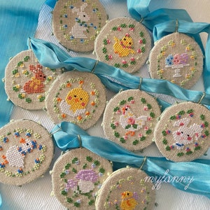 Könnte beinhalten: Neun runde, bestickte Ornamente mit verschiedenen Ostermotiven, darunter Hasen, Küken und Eier. Die Ornamente befinden sich auf einem hellen Hintergrund mit einem blauen Band.