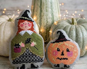 Pode incluir: Dois ornamentos de tecido acolchoados com desenhos em ponto cruz. Um é uma bruxa com cabelo ruivo, vestido verde e um gato preto. O outro é uma abóbora laranja redonda com um chapéu preto. Ambos têm verso de tecido cinza e botas pretas.