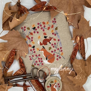 Può includere: Un disegno a punto croce con due volpi, bacche e foglie nei toni dell'arancione, rosso e verde. Il disegno è su tessuto beige, con un piccolo disegno rotondo di volpe e chiavi d'epoca. Il testo "myfanny" è visibile.
