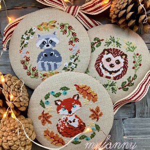 May include: Three round, beige, fabric ornaments with cross-stitch designs of a raccoon, hedgehog, and fox. Each ornament is surrounded by a wreath of leaves and berries. The ornaments are hanging from a red and white striped ribbon.