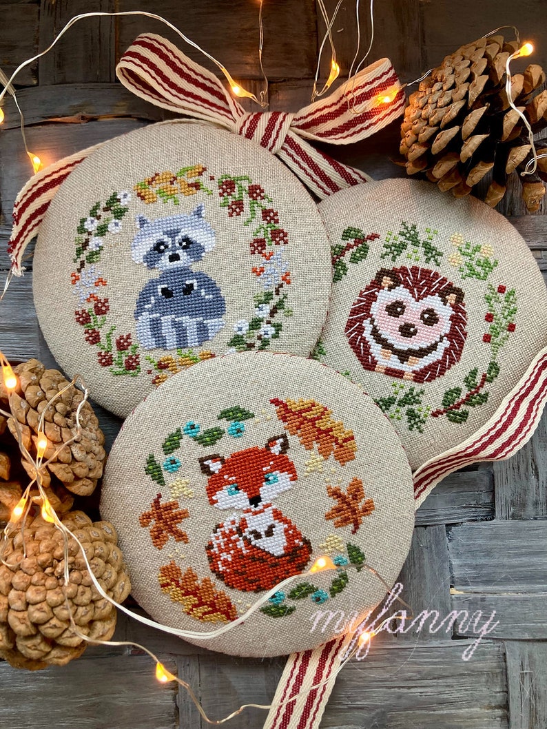 May include: Three round, beige, fabric ornaments with cross-stitch designs of a raccoon, hedgehog, and fox. Each ornament is surrounded by a wreath of leaves and berries. The ornaments are hanging from a red and white striped ribbon.