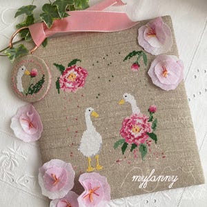 Può includere: Un ricamo a punto croce in lino beige con due oche bianche con becchi e zampe gialle e fiori rosa. Un punto croce circolare più piccolo con un'oca è attaccato con un nastro rosa. Il testo "myfanny" è scritto in corsivo. Sono attaccati fiori di stoffa rosa.