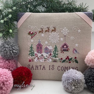 Può includere: Un ricamo a punto croce su un tessuto di lino beige con una scena invernale di case, pupazzi di neve e una slitta trainata da renne. Il testo "SANTA IS COMING" è ricamato sotto la scena. Il disegno è incorniciato da un nastro rosa.