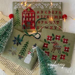 Può includere: Ornamenti natalizi a punto croce in tessuto verde oliva. I disegni includono una casa rossa, l'alfabeto, fiocchi di neve, alberi di Natale, un pupazzo di neve e una renna in una ghirlanda. Gli ornamenti sono decorati con filo rosso e verde.