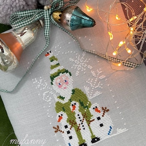 Könnte beinhalten: Ein Kreuzstichmuster eines grünen Zwergs mit weißem Bart, der mit zwei Schneemännern steht. Der Zwerg trägt einen grünen Hut mit weißem Besatz und einen grünen Mantel. Die Schneemänner tragen schwarze Hüte und Schals. Der Hintergrund ist ein weißer Stoff mit einem Schneeflockenmuster.