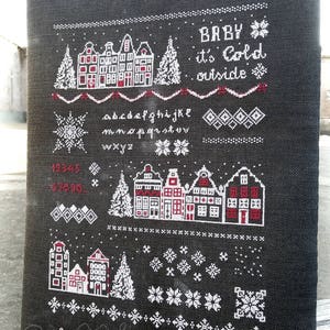 Può includere: Motivo a punto croce in bianco e nero con una scena invernale di case, alberi e fiocchi di neve. Il testo "BABY it's Cold outside" è ricamato in filo rosso.