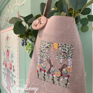 Può includere: Una borsa tote in lino beige con un ricamo a punto croce che raffigura due conigli in un giardino. Il disegno include un sole, fiori e alberi. La borsa ha un manico in pelle marrone e una piccola toppa rotonda ricamata a punto croce con un motivo di uccello.