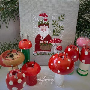 Può includere: Una collezione di tricotin in legno a forma di fungo con perni in metallo, caratterizzati da motivi a pois rossi e bianchi, accanto a un tessuto rettangolare grigio chiaro con un disegno a punto croce di un Babbo Natale con un cestino.