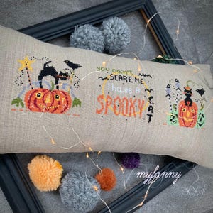 Può includere: Un cuscino beige a tema Halloween con disegni a punto croce. Il cuscino presenta una zucca, un gatto nero e il testo "You can't scare me, I have a spooky cat." Pompon decorativi e luci fatate circondano il cuscino.