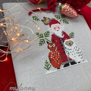 Può includere: Schema di ricamo a punto croce con Babbo Natale con due gufi, uno marrone e uno bianco, su uno sfondo di lino bianco. Lo schema è incorniciato da un bordo verde e fiocchi di neve.