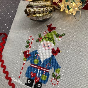 Può includere: Schema di punto croce di un allegro Babbo Natale che indossa un cappotto blu, un cappello verde e una barba bianca. Tiene un bastone di zucchero e un sacco di regali. Lo schema è su un tessuto di lino bianco.