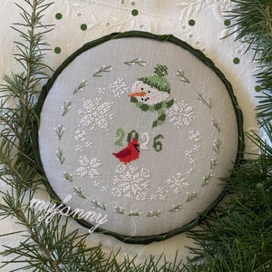 Può includere: Un disegno a punto croce rotondo e grigio con un pupazzo di neve con cappello e sciarpa verdi, un cardinale rosso e l'anno "2026". Il disegno è circondato da fiocchi di neve e verde, con un bordo verde.