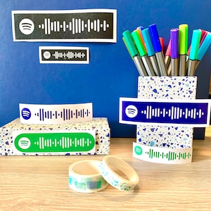 Peut inclure: Gros plan sur un bureau avec une variété de marqueurs colorés dans un gobelet à motif terrazzo. Il y a plusieurs autocollants blancs avec un logo Spotify noir et un code-barres. Il y a aussi deux rouleaux de ruban adhésif washi, l'un avec un motif bleu et blanc et l'autre avec un motif vert et bleu.