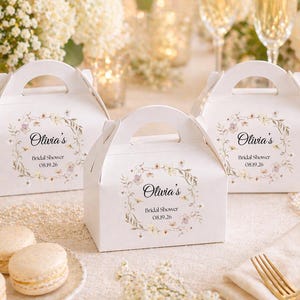Peut inclure: Trois boîtes à cadeaux blanches avec poignées, chacune ornée d'une couronne florale et du texte "Olivia's Bridal Shower 08.19.26." Les boîtes sont disposées sur une table avec des macarons, des fleurs et des flûtes à champagne.