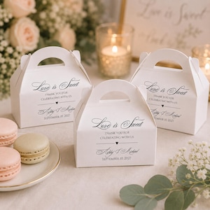 May include: Three white favor boxes with handles, each printed with "Love is Sweet" and personalized text. The boxes are arranged with macarons and flowers in the background, creating a celebratory atmosphere.