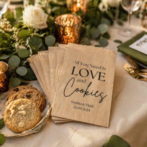 May include: Brown paper cookie bags with the text "All You Need Is LOVE and Cookies" in black script. The bags are on a table with cookies and floral decorations. The date "24.09.2024" is printed on the bags.