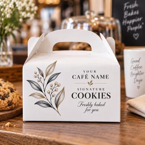 May include: A white cardboard bakery box with a handle, featuring a botanical illustration of leaves and berries on the left side. The text reads: YOUR CAFÉ NAME, SIGNATURE COOKIES, and Freshly baked for you in black serif and script fonts.