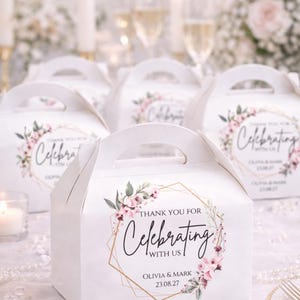 Peut inclure: Boîtes à cadeaux blanches avec poignées, décorées de motifs floraux et de cadres géométriques dorés. Les boîtes portent l'inscription "Thank You For Celebrating With Us" ainsi que des noms et une date. Bougies et coupes de champagne en arrière-plan.