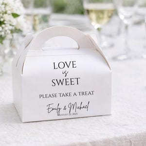 May include: White favor box with a handle, featuring the text "LOVE is SWEET PLEASE TAKE A TREAT" and names "Emily & Michael" with a date. The box is on a white tablecloth, with flowers and glasses in the background.