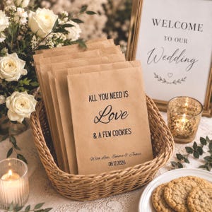 Puede incluir: Una cesta de mimbre contiene varias bolsas de papel marrón con el texto "ALL YOU NEED IS Love & A FEW COOKIES" impreso. Las bolsas son para una boda, con los nombres Emma & James y la fecha 06.12.2026 impresos en ellas. Un letrero que dice "WELCOME TO OUR Wedding" está en el fondo.