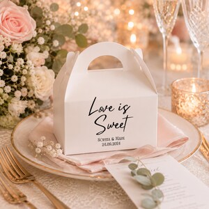 May include: A white cardboard favour box with a handle, featuring the black printed text Love is Sweet, Sophia & Mark, and the date 24.09.2024. The box has a smooth finish and a classic gable top design, presented as a decorative container.