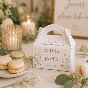 May include: A white wedding favour box with a handle, floral designs, and the names "Olivia and James" with the date "June 21, 2026." The box is on a table with macarons and a lit candle in the background.