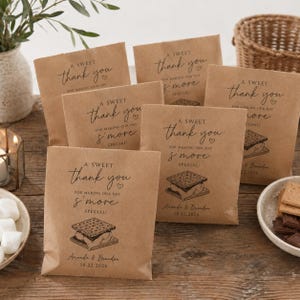 May include: Brown paper favor bags with a s'more illustration and the words "A Sweet Thank You." Each bag is printed with the names "Amanda & Brandon" and the date "10.22.2026." The bags are on a wooden table.
