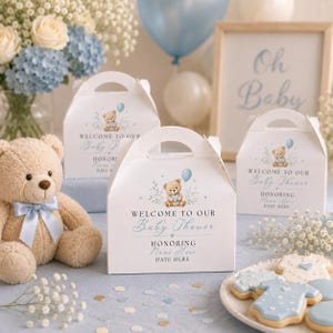 Może przedstawiać: Białe kartonowe pudełka na upominki z uchwytami, ozdobione ilustracją misia, niebieskim balonem i napisem Welcome to our Baby Shower. Obok znajduje się beżowy miś z niebieską wstążką oraz ciasteczka w kształcie dziecięcych body.