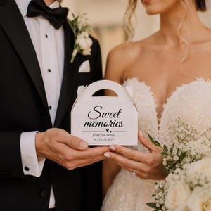 Puede incluir: Una caja de cartón blanca con asa, que presenta el texto impreso en negro Sweet memories, dos pequeños iconos de corazón y los nombres Jessica & Brian junto a la fecha August 14, 2024, centrados en la parte frontal.