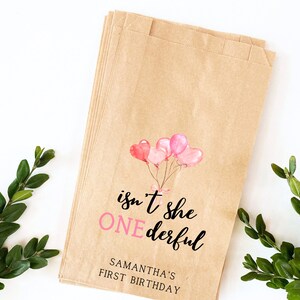 K&ouml;nnte beinhalten: Braune Papiert&uuml;ten mit einem Design aus rosa und wei&szlig;en Aquarellballons. Der Text auf der T&uuml;te lautet "isn't she ONEderful" und "SAMANTHA'S FIRST BIRTHDAY".