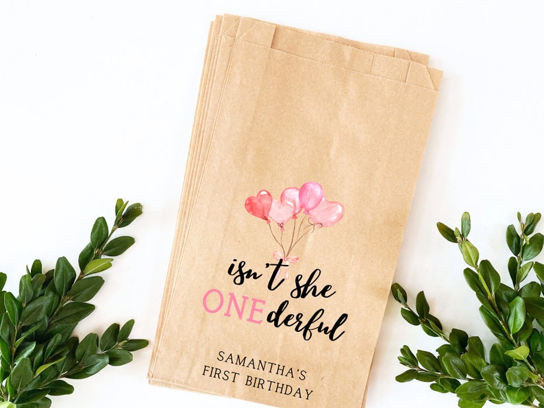 K&ouml;nnte beinhalten: Braune Papiert&uuml;ten mit einem Design aus rosa und wei&szlig;en Aquarellballons. Der Text auf der T&uuml;te lautet "isn't she ONEderful" und "SAMANTHA'S FIRST BIRTHDAY".