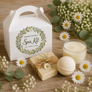 May include: A white gift box with a handle featuring a green botanical wreath and the text "PAMPER YOURSELF Spa Kit relax • refresh • recharge". Beside it sits a rectangular bar of soap wrapped in twine, a round bath bomb, and a glass candle.