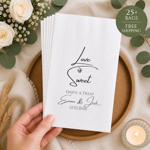 May include: A pile of white paper treat bags, each printed with the words "Love is Sweet" in black script. The bags are personalised with names and a date. The image also shows a wooden tray, flowers, and a lit candle.
