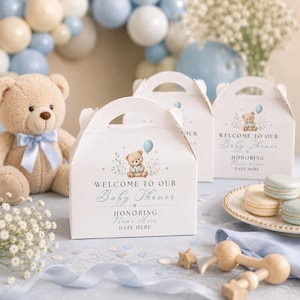 Può includere: Tre scatole regalo in cartone bianco con manici, ognuna con l'illustrazione di un orsacchiotto che tiene un cuore blu e un palloncino, con il testo Welcome to our Baby Shower, Honoring Name Here e Date Here stampato in blu.