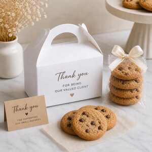 Op de afbeelding: Een witte geschenkdoos met een handvat en de tekst "Thank you" erop. Een stapel chocolate chip cookies is verpakt in doorzichtig cellofaan en vastgebonden met een strik. Een kleine kaart leest "Bedankt voor het steunen van ons kleine bedrijf."
