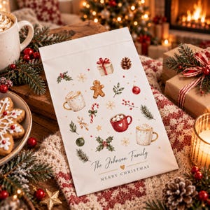 Peut inclure: Un sac en papier rectangulaire blanc orné d'un motif festif composé de bonshommes en pain d'épices, de sucres d'orge, de tasses de chocolat chaud, de pommes de pin et de houx. Le texte indique The Johnson Family et Merry Christmas.