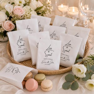 May include: White paper treat bags with the text "Love is Sweet" and the names "Emma & Jack" with a date. The bags are in a woven basket, surrounded by macarons and flowers, perfect for a wedding or party.