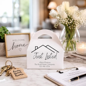 May include: A white cardboard gift box with a handle, featuring a black house icon and the text Just Listed, Olivia Thomas, The Real Estate Company, and 123-456-1234. The box is displayed alongside a wooden keychain, a sign, and paperwork.